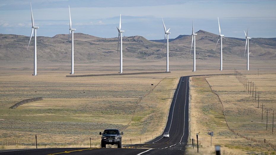 A pickup truck travels along a rural highway with wind turbines rising across the Wyoming plains.